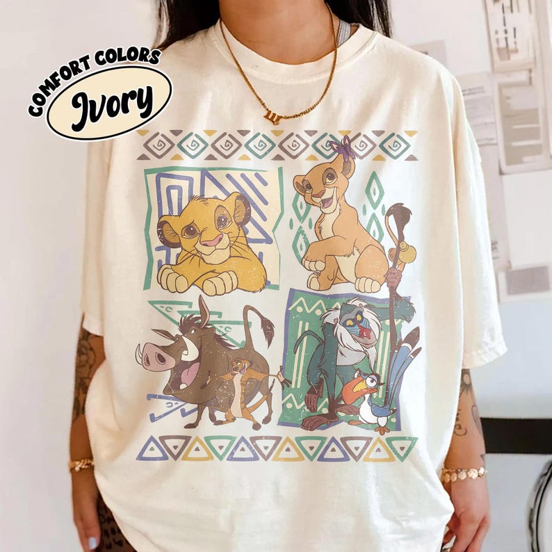 May include: Ivory-colored t-shirt featuring a colorful graphic of characters from The Lion King. The design includes Simba, Timon, Pumbaa, and Rafiki, with geometric patterns. The shirt has the words "Comfort Colors" and "Ivory" printed on it.