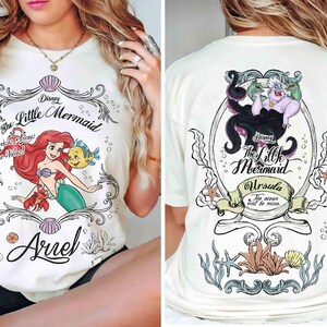 May include: Cream-coloured t-shirt with a Disney's The Little Mermaid graphic. The front shows Ariel, Flounder, and Sebastian, with text reading "The Little Mermaid" and "Ariel". The back features Ursula with the text "Ursula" and "The ocean will be mine."