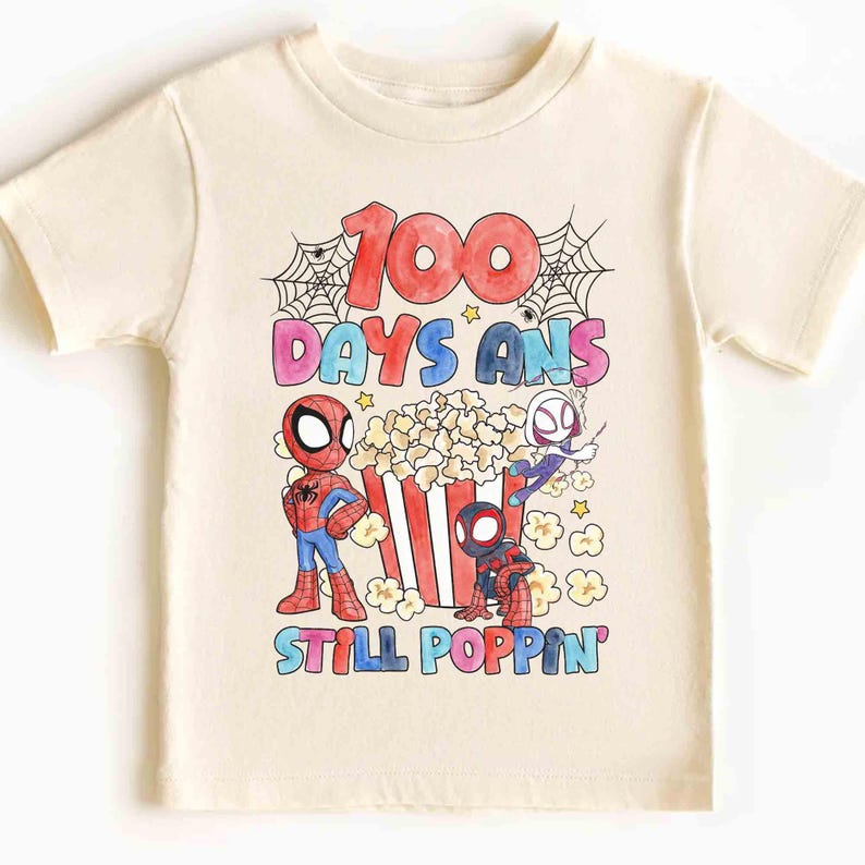 May include: Cream-colored t-shirt featuring a colorful graphic with the text "100 DAYS ANS STILL POPPIN". The design includes cartoon Spider-Man characters, popcorn, and spiderwebs. A fun and festive shirt for kids.