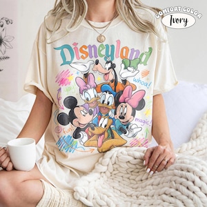 K&ouml;nnte beinhalten: Ein elfenbeinfarbenes T-Shirt mit einer bunten Grafik von Disney-Figuren, darunter Mickey Maus, Minnie Maus, Donald Duck, Daisy Duck, Goofy und Pluto. Der Text lautet Disneyland, whoa!, yay!, fun! und magic! mit krakeligen Akzenten.