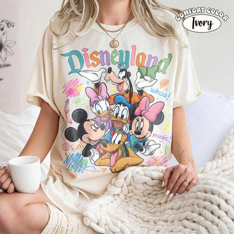 K&ouml;nnte beinhalten: Ein elfenbeinfarbenes T-Shirt mit einer bunten Grafik von Disney-Figuren, darunter Mickey Maus, Minnie Maus, Donald Duck, Daisy Duck, Goofy und Pluto. Der Text lautet Disneyland, whoa!, yay!, fun! und magic! mit krakeligen Akzenten.