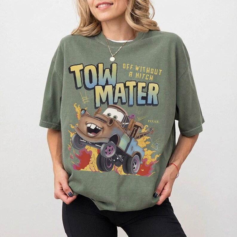 Może przedstawiać: Zielona koszulka z nadrukiem Złomka z filmu Auta studia Pixar. Projekt zawiera napis "TOW MATER OFF WITHOUT A HITCH" w postarzonej czcionce, z płomieniami i kresk&oacute;wkowym wizerunkiem Złomka.