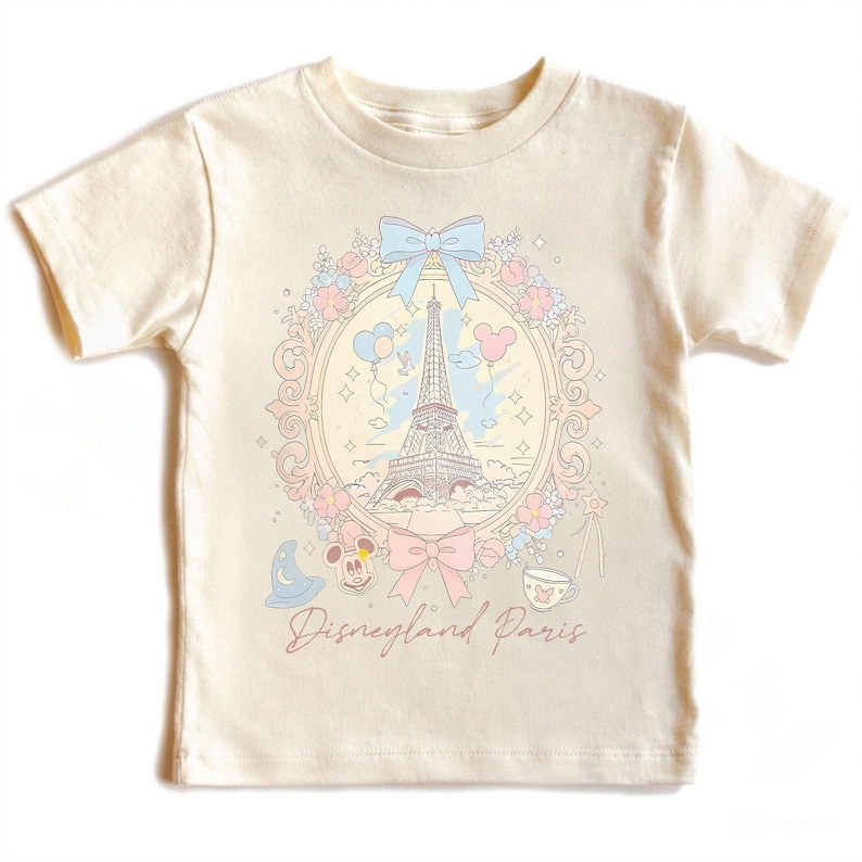 Peut inclure: T-shirt cr&egrave;me avec un motif Disneyland Paris. Le graphisme comprend la Tour Eiffel, Mickey Mouse et d'autres &eacute;l&eacute;ments fantaisistes dans un cadre d&eacute;coratif, avec le texte "Disneyland Paris".