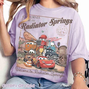 Op de afbeelding: Orchidee-kleurig T-shirt met een afbeelding van personages uit de film Cars, waaronder Lightning McQueen. Het ontwerp bevat de tekst "California est. 2012 Radiator Springs" en "Welcome to Radiator Springs."