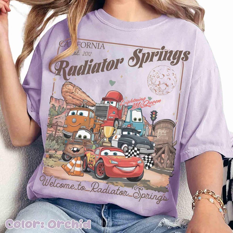Op de afbeelding: Orchidee-kleurig T-shirt met een afbeelding van personages uit de film Cars, waaronder Lightning McQueen. Het ontwerp bevat de tekst "California est. 2012 Radiator Springs" en "Welcome to Radiator Springs."
