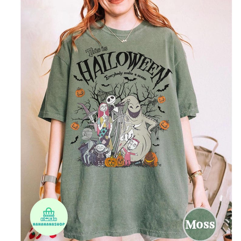May include: Moss green t-shirt featuring a Halloween-themed graphic. The design includes the text "This is Halloween" and various characters and pumpkins. The shirt is a comfortable, casual style.
