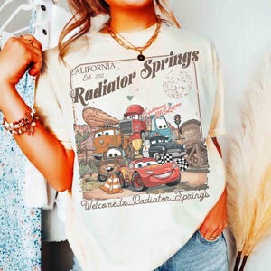 Op de afbeelding: Cr&egrave;mekleurig T-shirt met een afbeelding van personages uit de film Cars, waaronder Bliksem McQueen. Het ontwerp bevat de tekst "Radiator Springs" en "Welcome to Radiator Springs" tegen een woestijnlandschap.