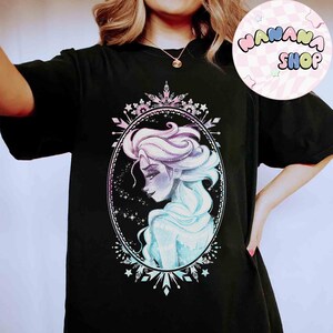 Op de afbeelding: Zwart T-shirt met een aquarel illustratie van een vrouw met vloeiend blauw en paars haar, omlijst in een ovaal met een decoratieve rand. Het ontwerp bevat sterren en sneeuwvlok accenten. Het shirt heeft ook de tekst "NANANA SHOP" in een cirkel.