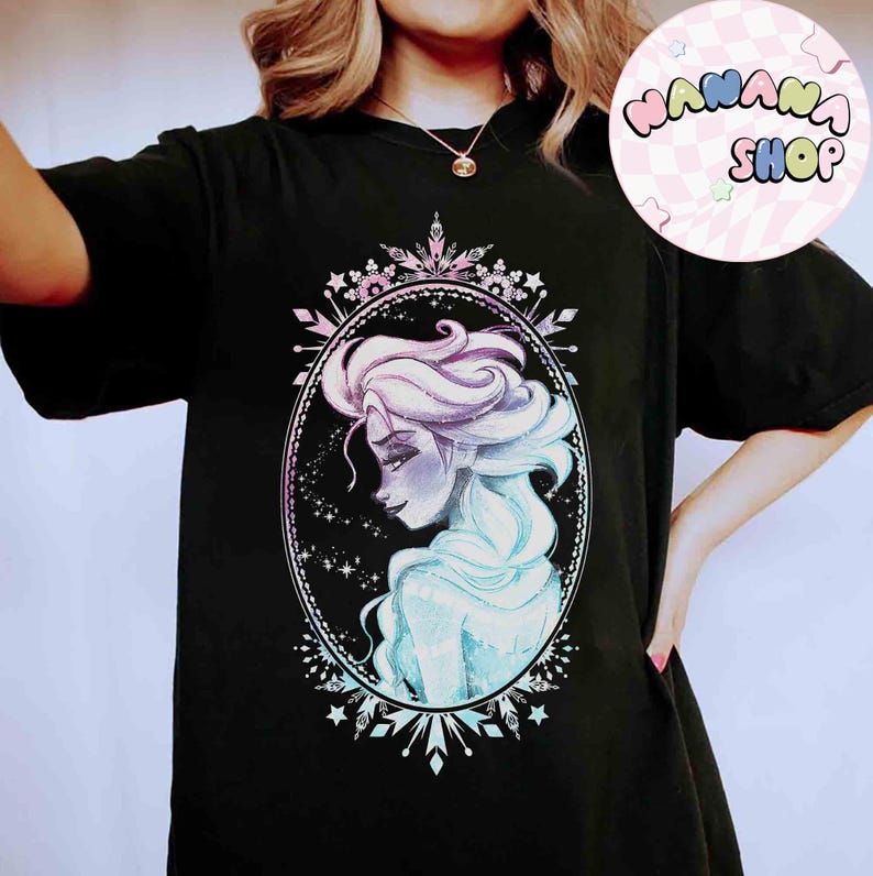 Op de afbeelding: Zwart T-shirt met een aquarel illustratie van een vrouw met vloeiend blauw en paars haar, omlijst in een ovaal met een decoratieve rand. Het ontwerp bevat sterren en sneeuwvlok accenten. Het shirt heeft ook de tekst "NANANA SHOP" in een cirkel.