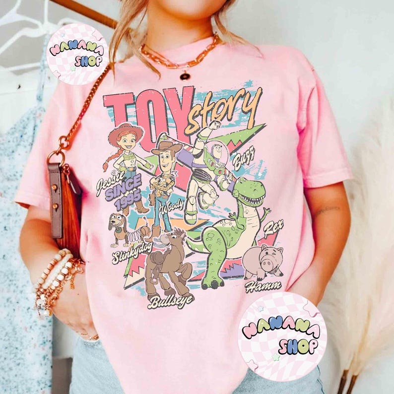 Puede incluir: Camiseta rosa con la frase "Toy Story" en una fuente retro, con los personajes de dibujos animados Jessie, Woody, Buzz Lightyear, Rex, Hamm, Slinky Dog y Bullseye. La camiseta tambi&eacute;n incluye el texto "Since 1995".