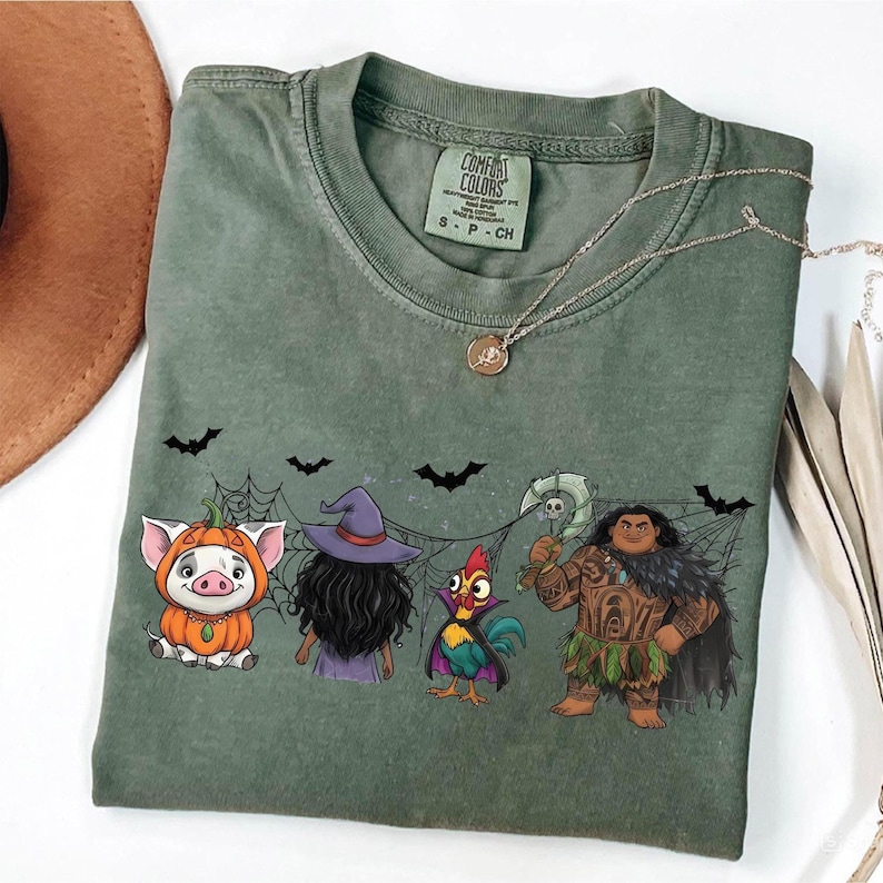 May include: Sage green Comfort Colors t-shirt featuring a Halloween-themed graphic. The design includes cartoon characters, bats, and spiderwebs. A gold necklace is draped across the shirt. The shirt is labeled S-P-CH.