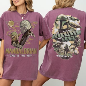 May include: A maroon t-shirt featuring a graphic of The Mandalorian and Grogu from Star Wars. The front design includes the text "MANDALORIAN" and "THIS IS THE WAY." The back design also features the text "THE MANDALORIAN AND GROGU."