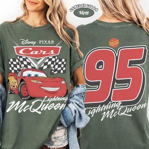 May include: A moss green t-shirt featuring Lightning McQueen from Disney Pixar's Cars. The front graphic includes the Cars logo, a checkered flag, and Lightning McQueen. The back displays a large number 95 and "Lightning McQueen" in white.