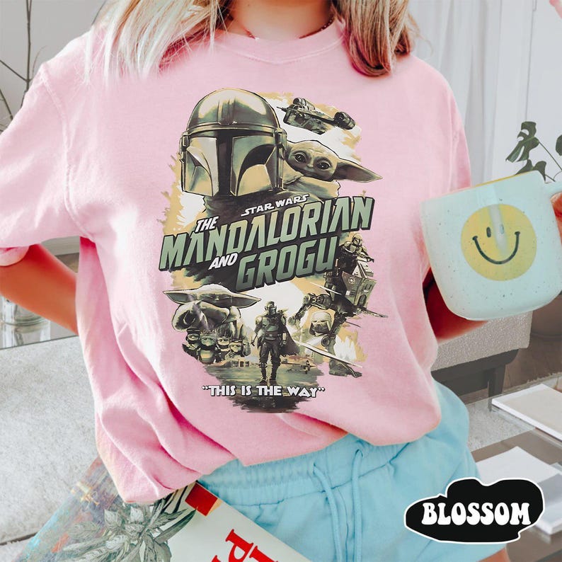 May include: Pink Star Wars "The Mandalorian and Grogu" graphic t-shirt. The design features the Mandalorian helmet, Grogu, and other characters with the phrase "This is the Way". The shirt is a light pink color.