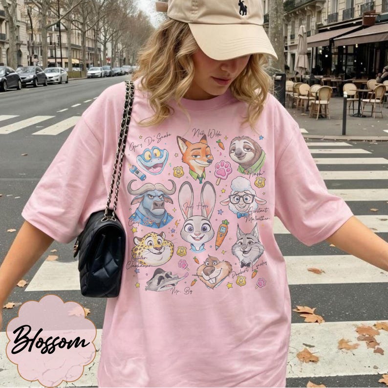 Pu&ograve; includere: T-shirt rosa chiaro con illustrazioni di personaggi di Zootropolis. La maglietta ha un girocollo e maniche corte. La parola "Blossom" &egrave; stampata in corsivo in basso a sinistra.