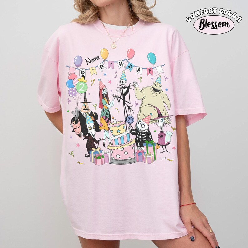Op de afbeelding: Een lichtroze t-shirt met een grafische print van personages uit The Nightmare Before Christmas die feesthoedjes dragen. Het ontwerp bevat een verjaardagstaart, ballonnen en cadeautjes, met de tekst "Name" en "Birthday" erboven gedrukt.