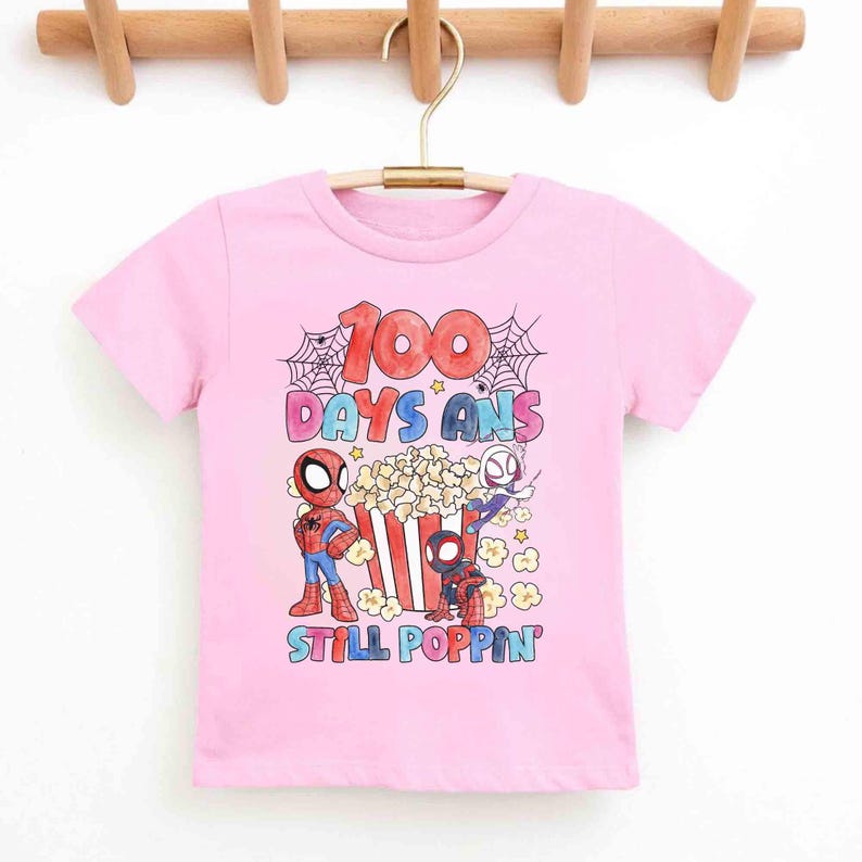 May include: Pink t-shirt featuring Spider-Man characters and popcorn. The design includes the text "100 DAYS ANS STILL POPPIN" in colorful letters, with spiderwebs and stars. The shirt is hanging on a wooden hanger.