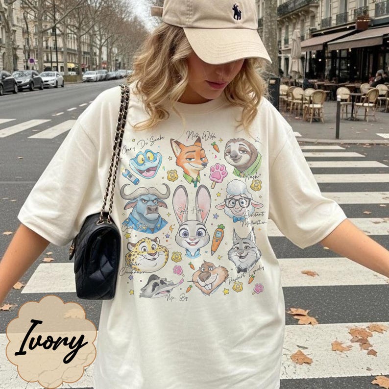 Pu&ograve; includere: T-shirt color avorio con una grafica colorata di personaggi di animali dei cartoni animati. La maglietta ha un girocollo e maniche corte. La parola "Ivory" &egrave; stampata in basso sulla maglietta.