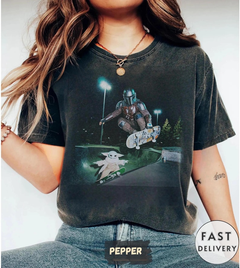 May include: Dark gray t-shirt featuring a graphic of The Mandalorian and Grogu skateboarding in a skate park. The design includes a detailed illustration of the characters and skateboards, set against a night scene with lights. The shirt has a vintage, worn look.