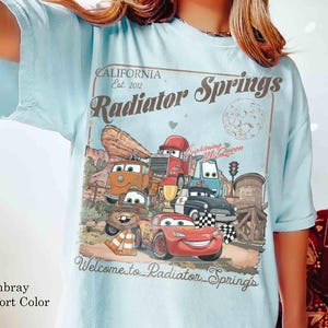 Op de afbeelding: Lichtblauw T-shirt met een afbeelding van de personages uit de film Cars, waaronder Lightning McQueen, en de tekst "Radiator Springs". Het ontwerp bevat een woestijnlandschap en de woorden "California Est. 2012."
