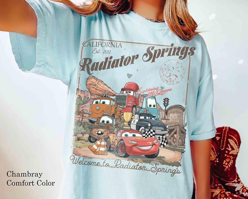Op de afbeelding: Lichtblauw T-shirt met een afbeelding van de personages uit de film Cars, waaronder Lightning McQueen, en de tekst "Radiator Springs". Het ontwerp bevat een woestijnlandschap en de woorden "California Est. 2012."