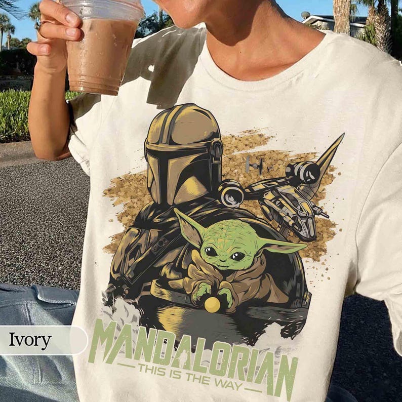 Puede incluir: Camiseta de color marfil con un dise&ntilde;o de The Mandalorian y Grogu. El dise&ntilde;o incluye una ilustraci&oacute;n detallada del casco del Mandaloriano, Grogu y una nave espacial, con el texto "MANDALORIAN - THIS IS THE WAY".