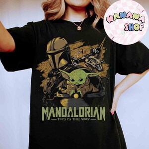 Puede incluir: Camiseta negra con un dise&ntilde;o de The Mandalorian y Grogu, una nave espacial y el texto "MANDALORIAN - THIS IS THE WAY" en verde y dorado. El dise&ntilde;o est&aacute; sobre un fondo texturizado, con el logo "NANANA SHOP" en la esquina superior derecha.