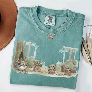 May include: A teal Comfort Colors t-shirt featuring a graphic of a character from a popular science fiction series. The design shows the character in a forest setting with the word "Coo!" printed on the shirt. A silver necklace with a pendant is on the shirt.