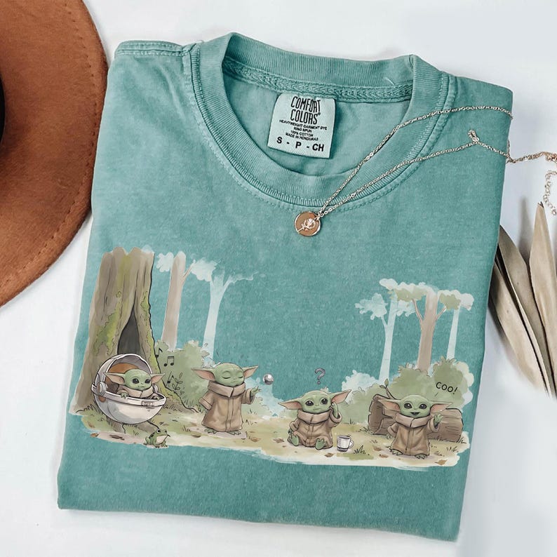 May include: A teal Comfort Colors t-shirt featuring a graphic of a character from a popular science fiction series. The design shows the character in a forest setting with the word "Coo!" printed on the shirt. A silver necklace with a pendant is on the shirt.
