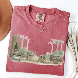 May include: A red Comfort Colors t-shirt featuring a graphic of Grogu from The Mandalorian. The design shows Grogu in various poses in a forest setting. A silver necklace with a pendant is draped across the shirt.