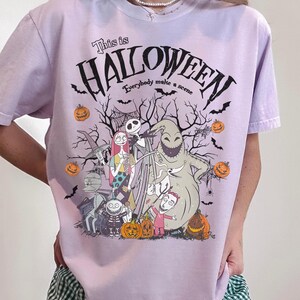May include: A light purple t-shirt featuring a Halloween-themed graphic. The design includes the text "This is Halloween" and "Everybody make a scene" above a group of cartoon characters, pumpkins, and bats.
