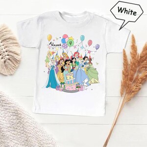 May include: White t-shirt featuring a colorful graphic of cartoon princesses celebrating a birthday. The design includes a birthday cake, balloons, and the number 4. The word "Name" is also visible.