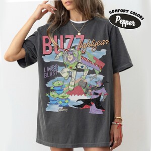 May include: Dark gray t-shirt featuring a graphic of Buzz Lightyear from Toy Story. The design includes the text "Buzz Lightyear", "Laser Blast", and "Aliens", along with illustrations of the characters. The shirt is labeled "Comfort Colors Pepper".