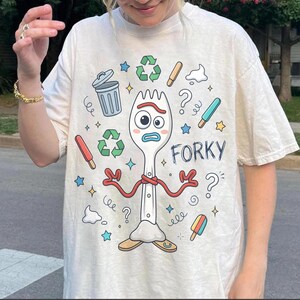 K&ouml;nnte beinhalten: Cremefarbenes T-Shirt mit einem Cartoon-Gabel-Charakter namens Forky aus Toy Story. Das Design umfasst Recycling-Symbole, Fragezeichen und bunte Eis am Stiel. Das Wort "Forky" ist in Blau gedruckt.