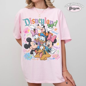 K&ouml;nnte beinhalten: Ein hellrosa T-Shirt mit einer bunten Grafik von Mickey Maus, Minnie Maus, Donald Duck, Daisy Duck, Goofy und Pluto. Der Schriftzug "Disneyland" steht oben, w&auml;hrend "yay!", "whoa!", "fun!" und "magic!" um die Figuren herum verteilt sind.