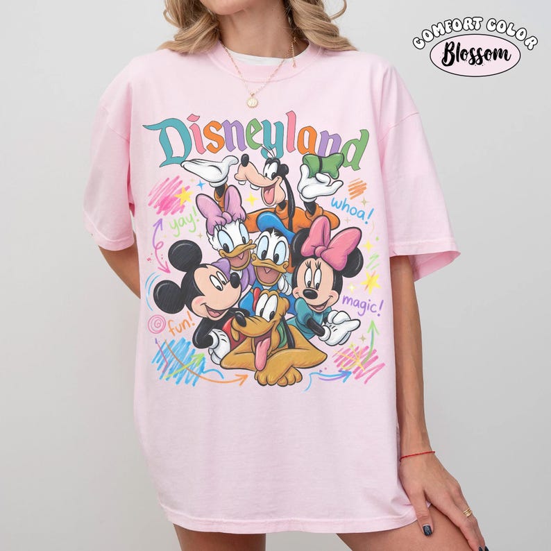K&ouml;nnte beinhalten: Ein hellrosa T-Shirt mit einer bunten Grafik von Mickey Maus, Minnie Maus, Donald Duck, Daisy Duck, Goofy und Pluto. Der Schriftzug "Disneyland" steht oben, w&auml;hrend "yay!", "whoa!", "fun!" und "magic!" um die Figuren herum verteilt sind.