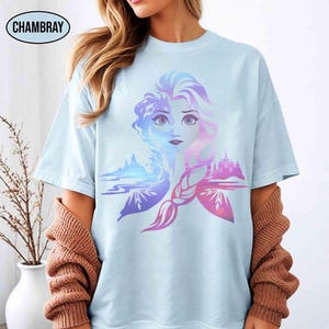 May include: Light blue t-shirt featuring a colorful graphic of a woman's face with a braided hairstyle, and a landscape design. The word "CHAMBRAY" is printed on the top left. The design incorporates shades of blue, purple, and pink.