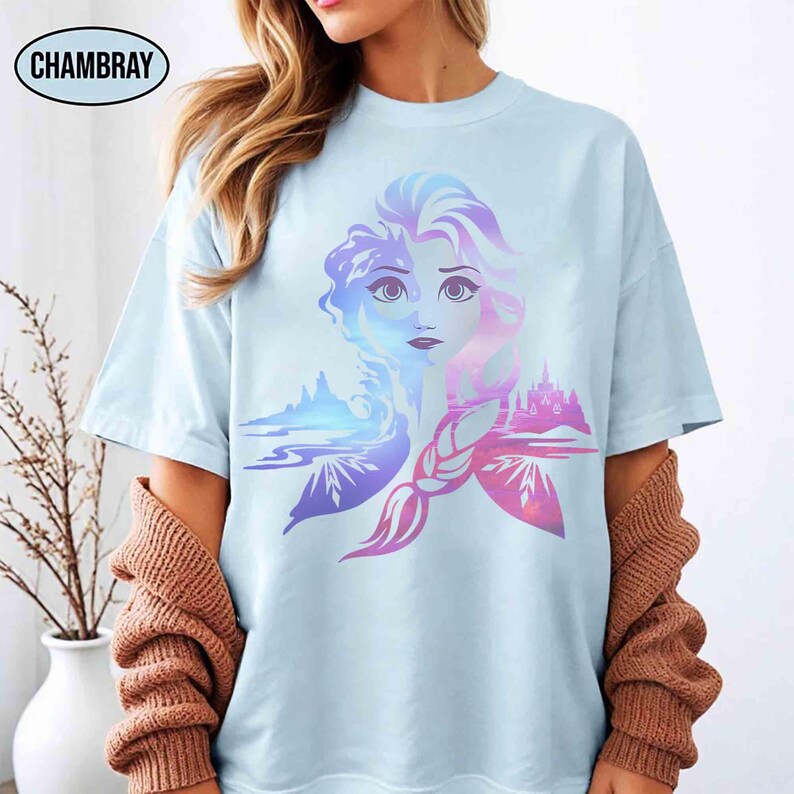 May include: Light blue t-shirt featuring a colorful graphic of a woman's face with a braided hairstyle, and a landscape design. The word "CHAMBRAY" is printed on the top left. The design incorporates shades of blue, purple, and pink.