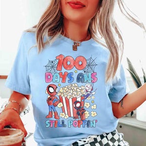 May include: Light blue t-shirt featuring a colorful graphic with the text "100 DAYS ANS STILL POPPIN'". The design includes cartoon Spider-Man characters, popcorn, and spiderwebs. The shirt is a short-sleeved crew neck.