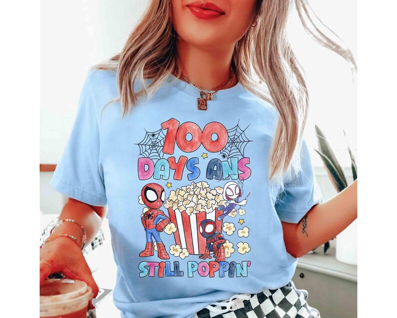 May include: Light blue t-shirt featuring a colorful graphic with the text "100 DAYS ANS STILL POPPIN'". The design includes cartoon Spider-Man characters, popcorn, and spiderwebs. The shirt is a short-sleeved crew neck.