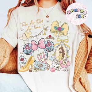 May include: Cream-colored t-shirt featuring a colorful graphic with the text "Tale As Old As Time." The design includes a pink bow, a yellow high heel, a rose in a glass dome, and a portrait of a woman in a yellow dress.