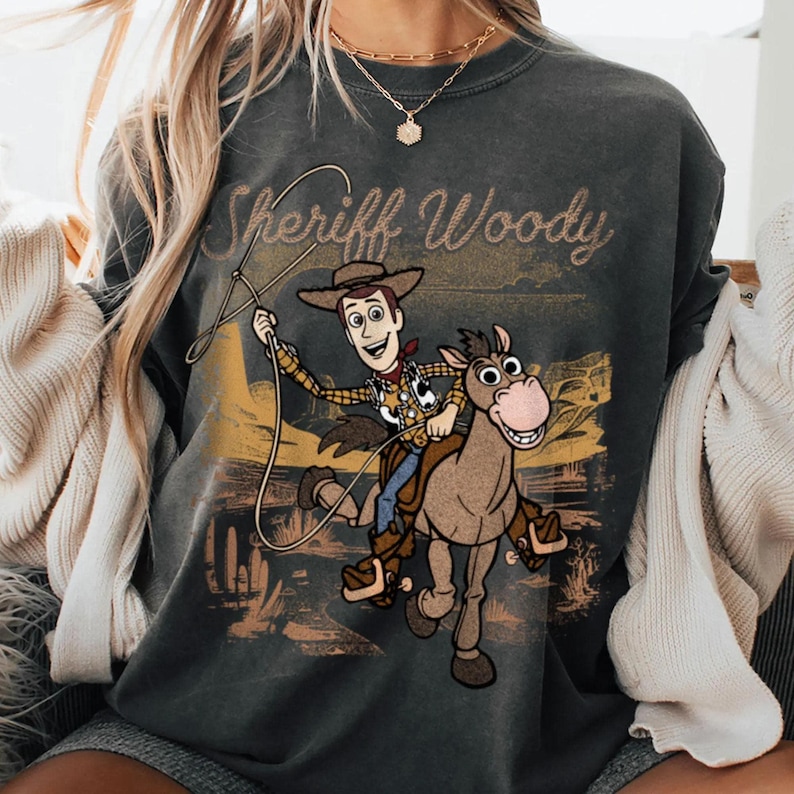 May include: Dark gray sweatshirt featuring Sheriff Woody from Toy Story riding a horse. The graphic includes the text "Sheriff Woody" in a distressed font, with a desert landscape background. The design is printed on a crew neck sweatshirt.