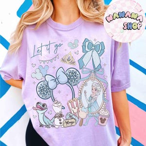 Może przedstawiać: Jasnofioletowa koszulka z nadrukiem graficznym z napisem "Let it go". Projekt zawiera ilustracje tiary, płatk&oacute;w śniegu, kokardy i portretu księżniczki. Widoczne są r&oacute;wnież postacie z kresk&oacute;wek i nap&oacute;j w kształcie Myszki Miki.