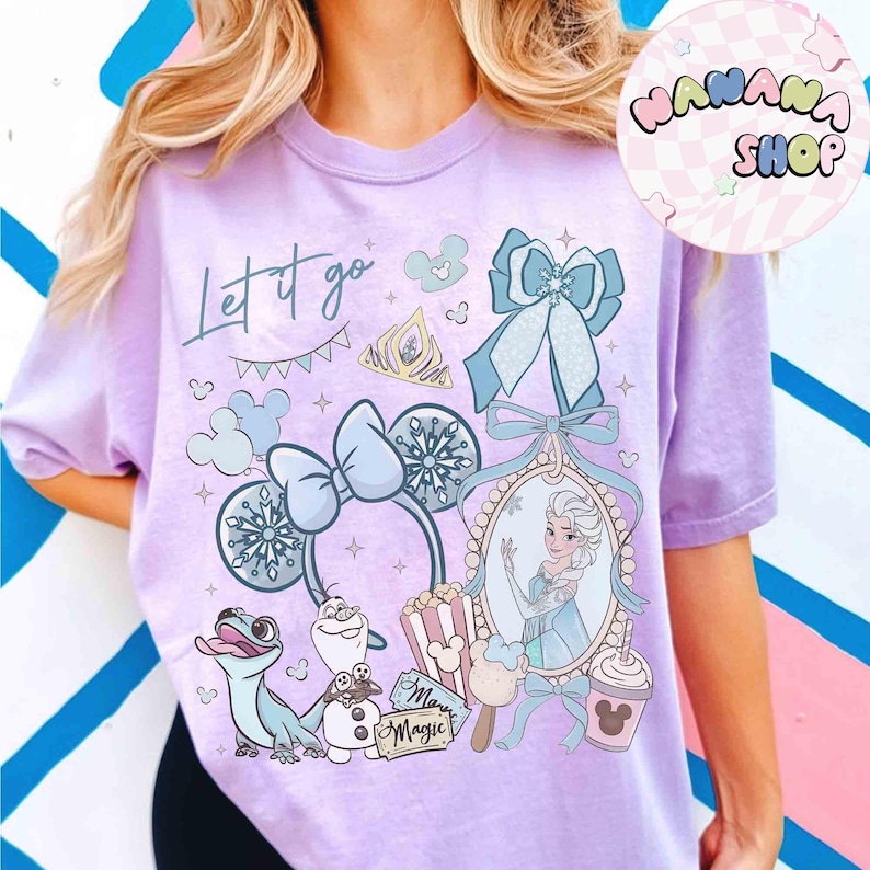 Może przedstawiać: Jasnofioletowa koszulka z nadrukiem graficznym z napisem "Let it go". Projekt zawiera ilustracje tiary, płatk&oacute;w śniegu, kokardy i portretu księżniczki. Widoczne są r&oacute;wnież postacie z kresk&oacute;wek i nap&oacute;j w kształcie Myszki Miki.
