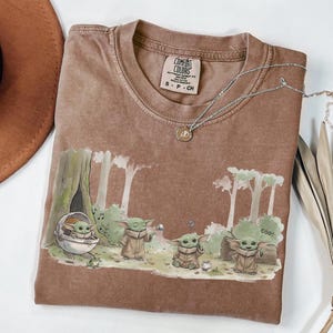 May include: Brown Comfort Colors t-shirt featuring a graphic of a green alien character in a forest scene. The shirt has a crew neck and short sleeves. A silver necklace with a pendant is draped across the shirt.