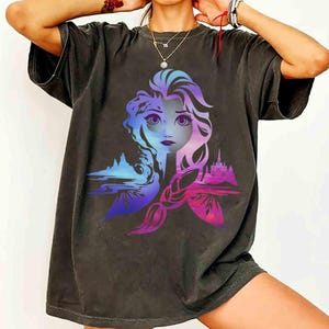 May include: Dark gray t-shirt featuring a colorful graphic of a woman's face with blue, purple, and pink hues. The design includes a castle and landscape elements, creating a silhouette-style image.