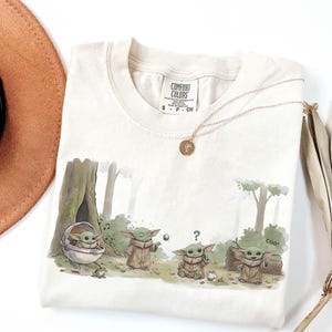May include: Cream-colored t-shirt featuring a cartoon illustration of a character in a forest setting. The graphic shows the character in different poses, with the word "cool" written on the shirt. A gold necklace with a pendant is on the shirt.