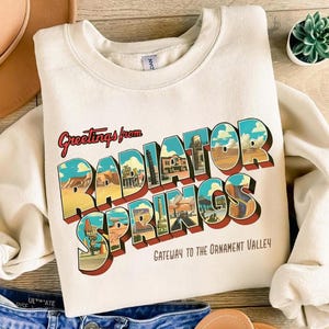 May include: Cream-colored sweatshirt featuring a colorful graphic that reads "Greetings from Radiator Springs" in a vintage postcard style. The design includes images of buildings and landscapes, with the text "Gateway to the Ornament Valley" below.