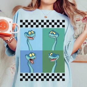 May include: Pale blue t-shirt with a graphic of four cartoon snakes, each with a different expression. The snakes are blue with yellow eyes, set against blue and green backgrounds, framed by a black and white checkered pattern.
