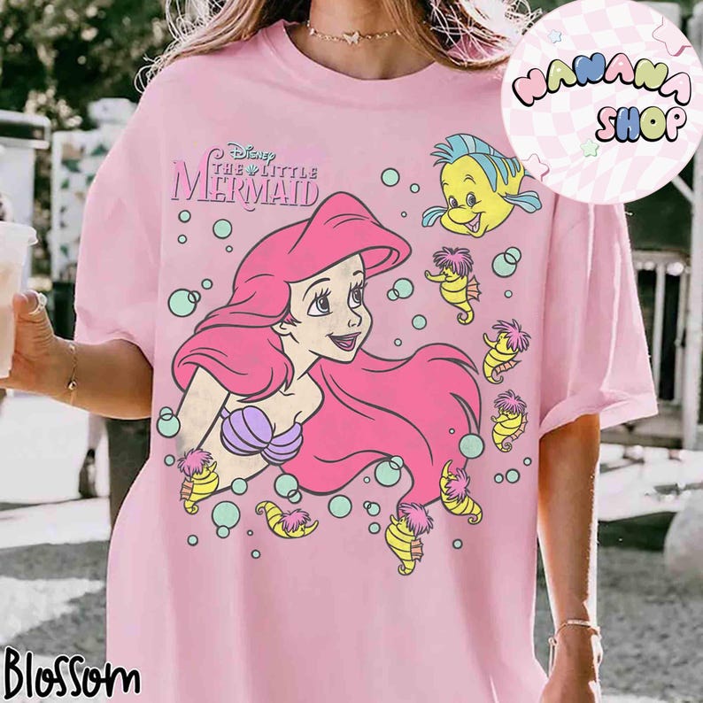 May include: Pink t-shirt featuring a graphic of Ariel from Disney's The Little Mermaid. The design includes Ariel with pink hair, a purple seashell bra, and Flounder. The shirt also has seahorses and bubbles.